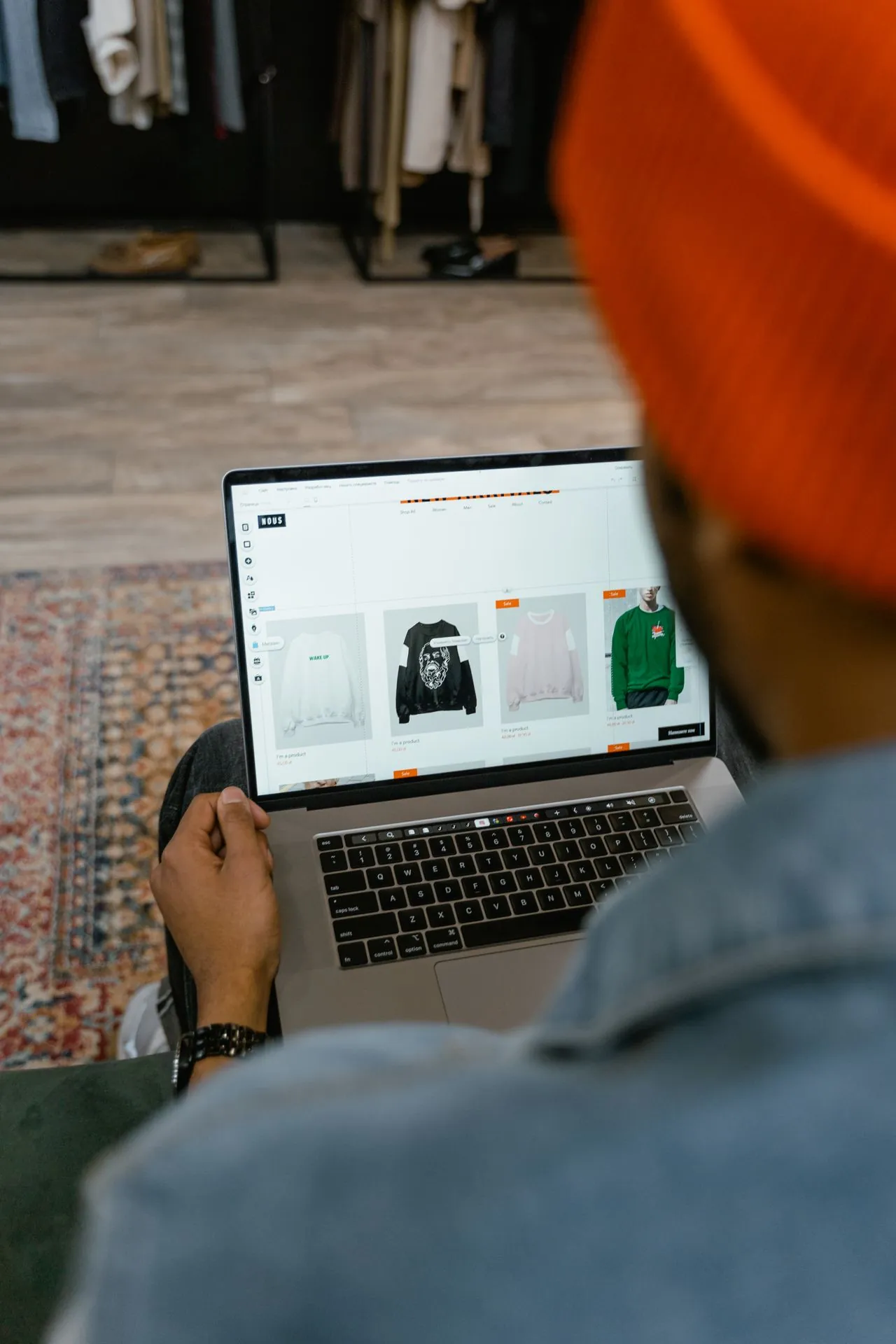 A store owner in an orange beanie edits his online store on a laptop in front of the clothing racks in his shop.