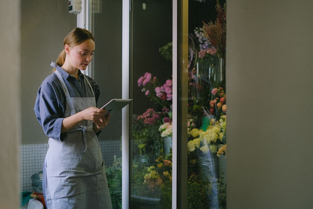 Una florista con delantal sostiene una tableta frente a una vitrina refrigerada llena de ramos de flores