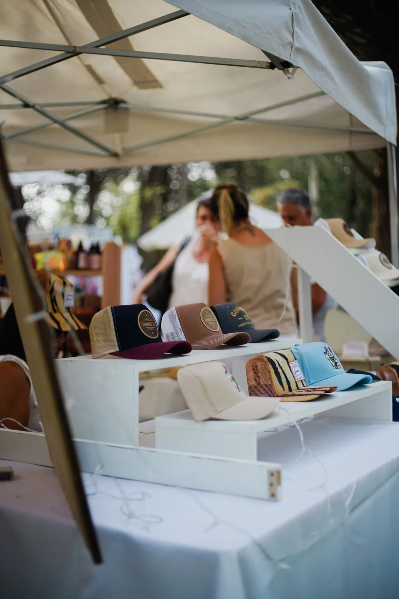 A market stall under a white tent: embroidered caps displayed on white stands, with customers chatting in the background