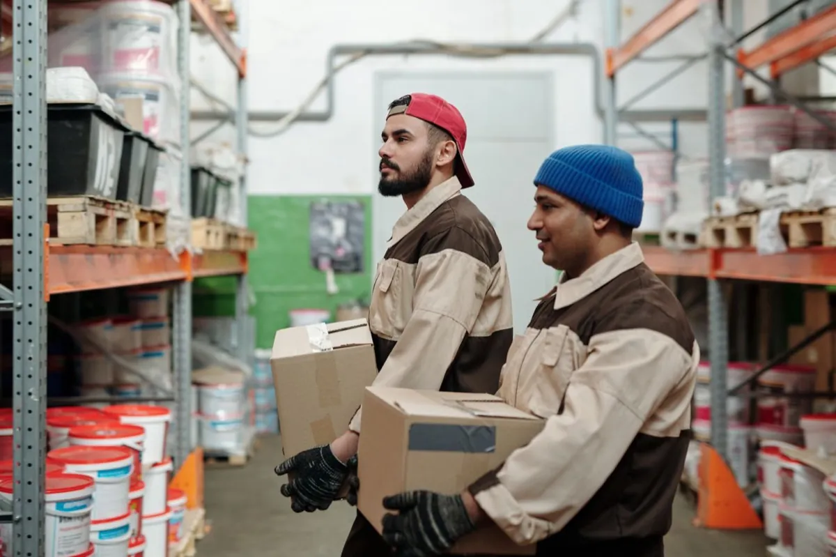 Deux employés portent des cartons dans l'entrepôt d'un fournisseur, entre des rayonnages de stock